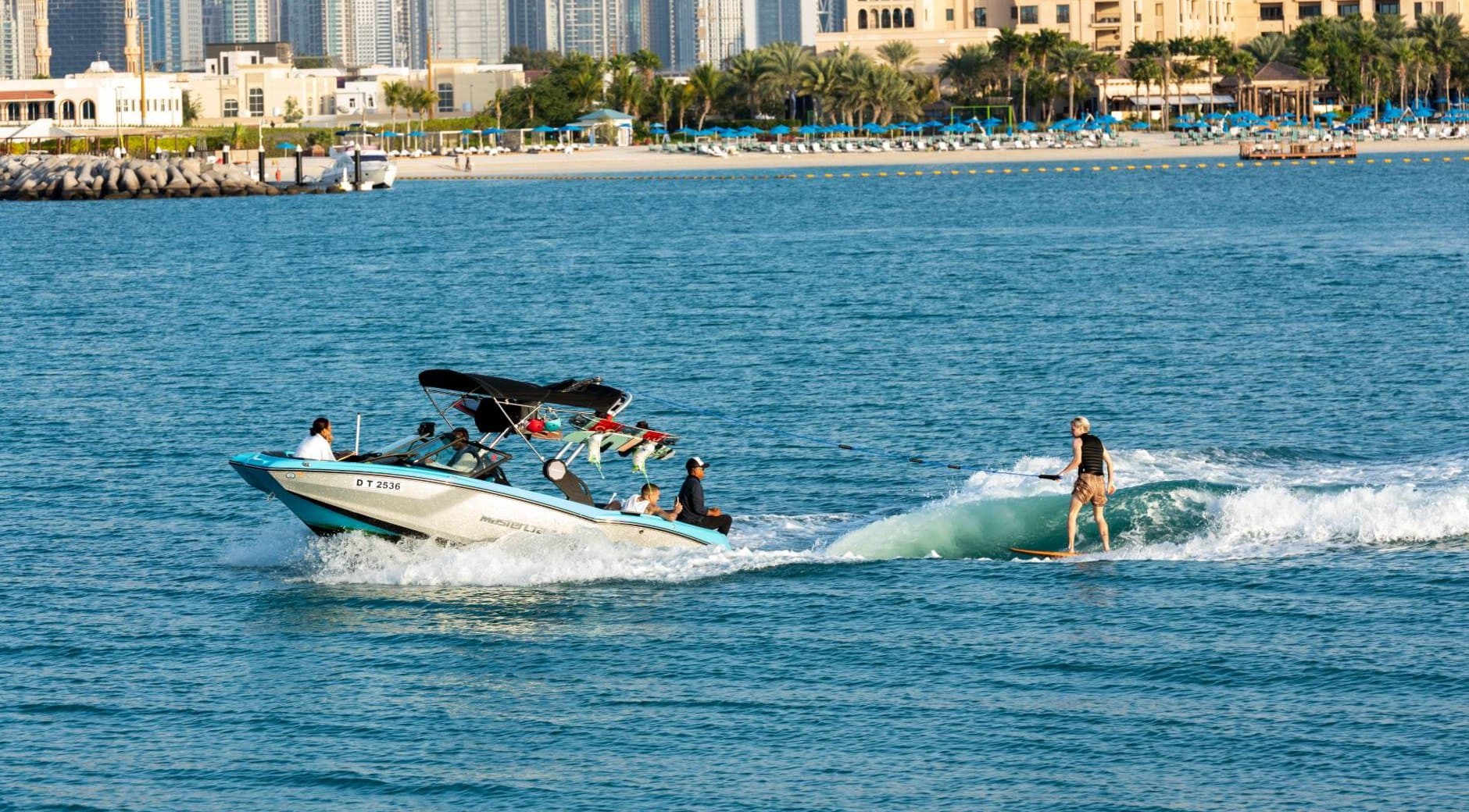 Вейксёрфер катается на волне за катером MasterCraft у побережья Джумейры, позади пляж, пальмы и небоскрёбы Дубая